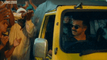 a man wearing sunglasses is driving a yellow car in front of a crowd of people with the year 2005 on the bottom right corner