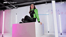 a woman in a green and black outfit is sitting on a cube in front of a sign that says academy