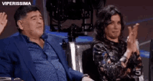 a man in a suit and a woman in a floral shirt are sitting next to each other and clapping .