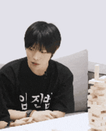 a man wearing a black shirt with korean writing on it sits at a table with a stack of wooden blocks