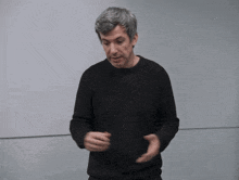 a man wearing a black sweater stands in front of a white wall