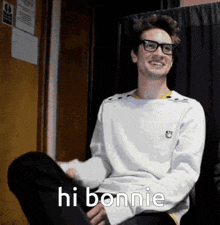 a young man wearing glasses and a white sweater says hi bonnie while sitting down