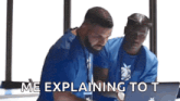 two men are sitting in front of a laptop computer and one of them is explaining to the other .