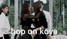 two women are hugging each other in front of a group of people and the words `` hop on koya '' are on the bottom .