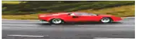 a red car is driving down a road with a grassy hill in the background