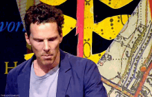 a man in a blue jacket and grey shirt is standing in front of a map with the letters h on it .