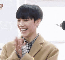 a young man wearing a plaid shirt and a tan coat is smiling and clapping .