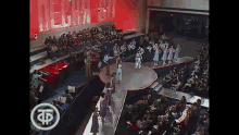 a group of people are standing on a stage in front of a crowd with the letter k on the bottom right