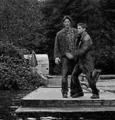 two men are standing on a dock next to a river .