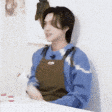a young man wearing a blue sweater and an apron is sitting at a table .