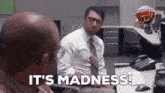a man sitting at a desk says it 's madness while another man looks on