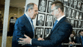 two men shaking hands in front of a wall with pictures of men
