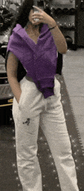 a woman in a purple shirt and white sweatpants is taking a selfie in a gym