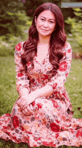 a woman in a floral dress sits in the grass with her hands folded