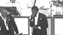 a man in a tuxedo sings into a microphone while another man stands behind him playing a keyboard