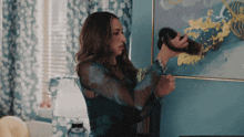 a woman in a blue shirt is holding a black object in front of a painting on a wall