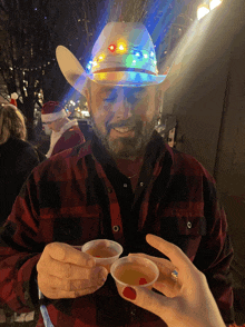 a man wearing a cowboy hat with lights on it holds two cups of liquid