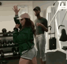 a man and a woman are standing in a gym . the woman is wearing a green jacket and shorts .