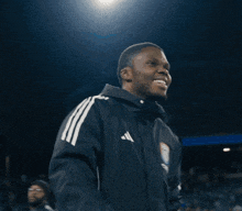 a man in an adidas jacket smiles while standing next to another man in a white shirt