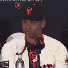 a man wearing a san francisco giants hat stands in front of a microphone holding a bottle of water