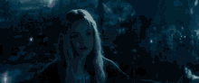 a woman with long blonde hair is standing in a dark forest surrounded by purple lights .