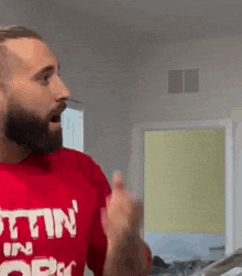 a man with a beard wearing a red t-shirt is standing in a room and making a funny face .