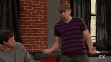 two men are sitting on a couch in a living room . one of the men is wearing a striped shirt .