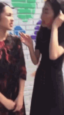 two women are standing next to each other and talking in front of a brick wall with graffiti on it .