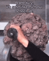 a person is holding a large rock in front of a sign that says warte kurz ich schmink mich nur schnell ab