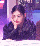 a woman in a leather jacket is laying on a bed with her hand on her chin .