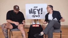 two men sitting in front of a sign that says hey
