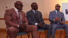 three men in suits are sitting in chairs and one has sunglasses on