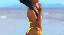 a woman in a yellow bikini is standing on a beach and touching her face .