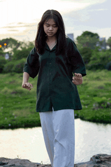 a woman in a green shirt and white pants stands in front of a river