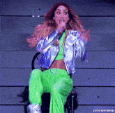a woman in green pants and a silver jacket is sitting on a chair and making a face