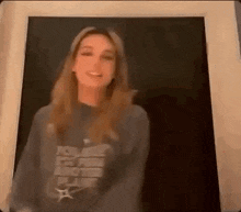 a woman in a gray sweatshirt is standing in front of a black wall and smiling .