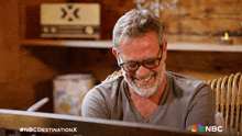 a man wearing glasses and a grey shirt is laughing with the nbc destination x logo behind him