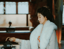 a woman wearing a white fur collar is pouring tea into a black teapot