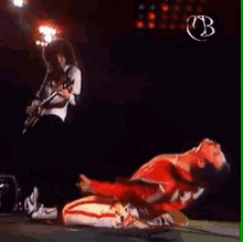 a man in a red jacket is laying on the ground on a stage while a man plays a guitar in the background .