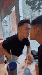 two men are sitting at a table with drinks and straws and one of them is blowing a kiss