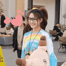 a woman wearing glasses and a tie dye shirt is holding a stuffed animal and smiling