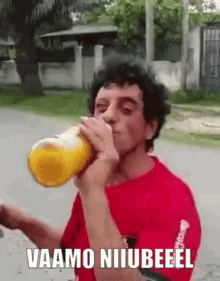 a man in a red shirt is drinking from a bottle with the words vaamo niiubeeel written on it .