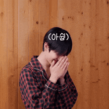 a man in a plaid shirt covering his face with his hands in front of a wood wall