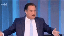 a man in a suit and tie is sitting at a table with his arms outstretched on a television show .