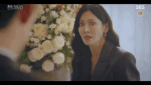 a woman in a black jacket is making a funny face while standing next to a man in front of a flower arrangement .