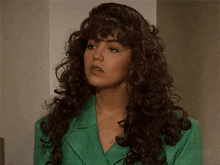 a woman with curly hair and a green jacket is standing in front of a wall .