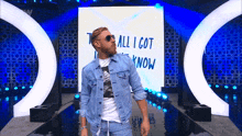 a man in a denim jacket and sunglasses stands in front of a screen that says " all i got now "