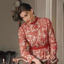 a woman wearing a red and white floral dress leans on a chair