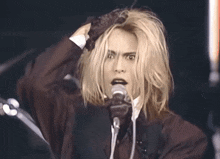 a man with blonde hair singing into a microphone with his hand on his head