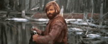 a man with a beard is standing next to a river holding a gun